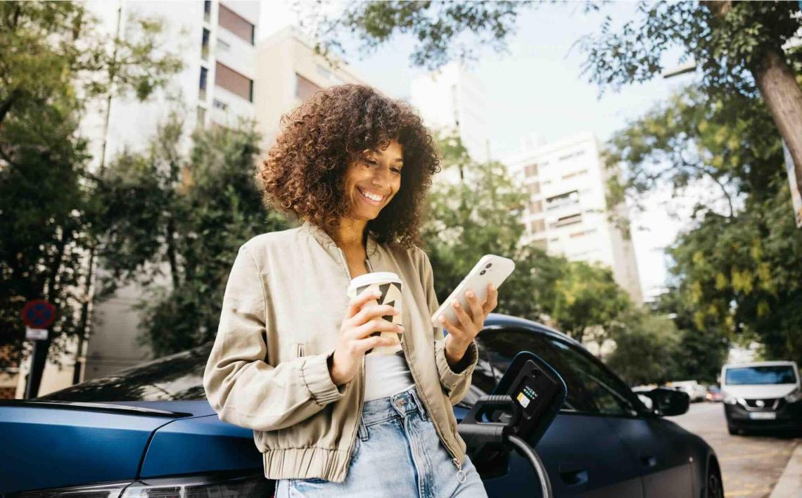Femme adossée à l’arrière d’une voiture bleue, souriante et regardant son téléphone.